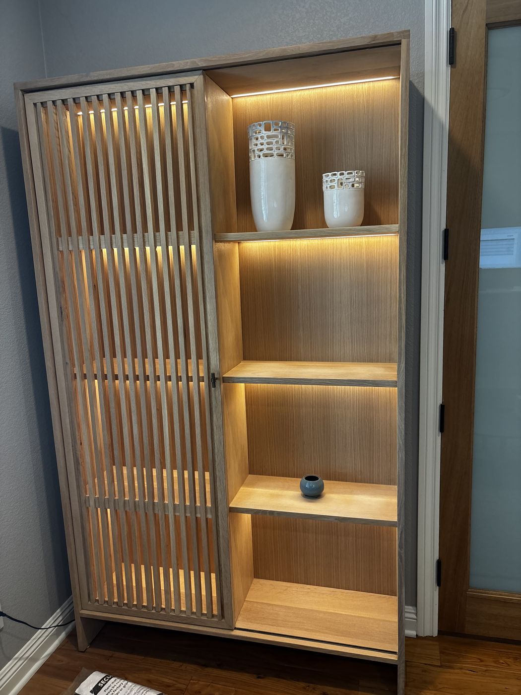 White oak display cabinet with a sliding slatted door and lit shelves