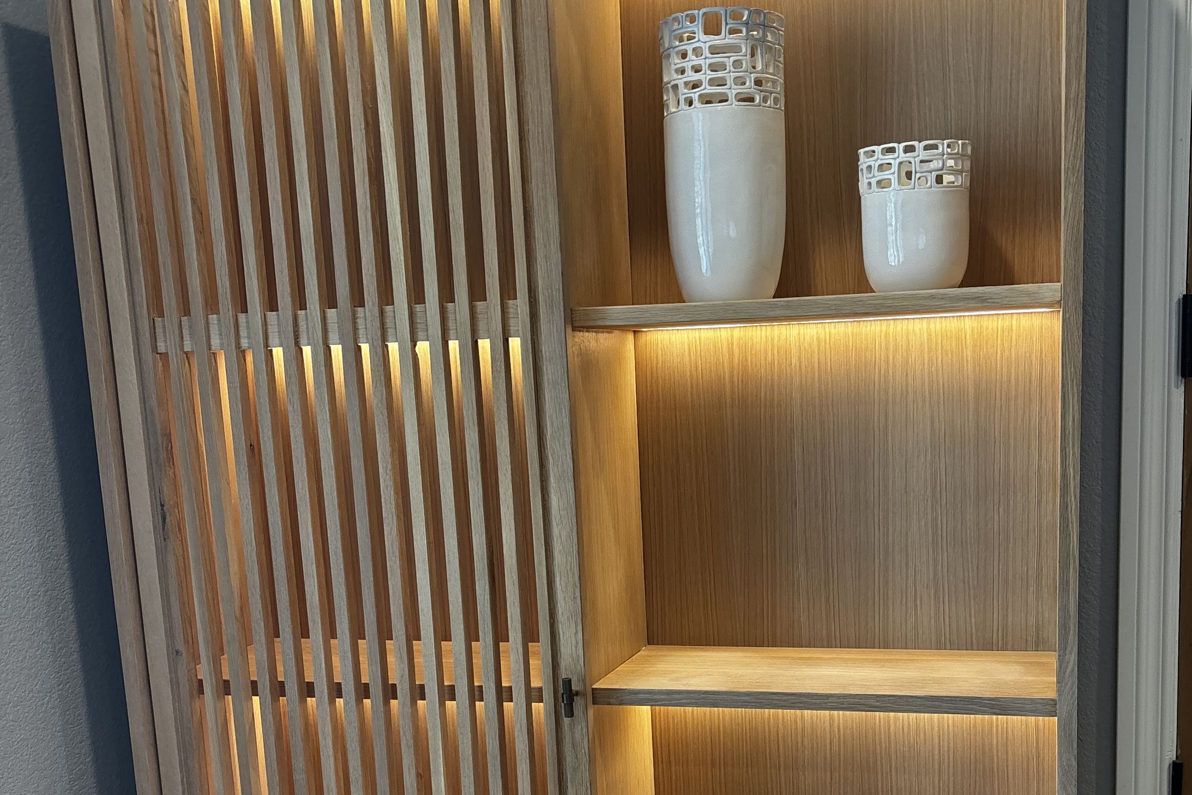 The Torrey Cabinet: a white oak cabinet with a sliding slatted door and softly lit shelves holding white ceramic vases